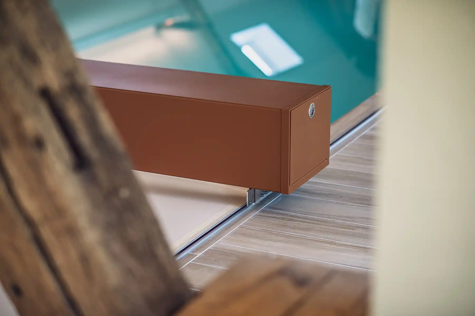 Boîtier rectangulaire marron fixé sur un rail en métal, posé sur un sol en bois clair, avec une piscine en arrière-plan.