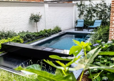 Petite piscine rectangulaire entourée de verdure, avec deux chaises longues bleues sur une terrasse pavée, près d'un mur blanc.