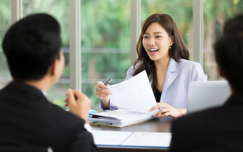 Une femme en blazer bleu clair tient un document et sourit à deux personnes assises de dos, autour d'une table en bois.