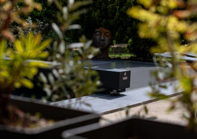 Une couverture de piscine grise est entourée de végétation, avec une sculpture de tête en arrière-plan.