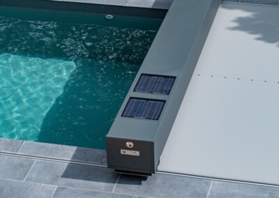 Travelhouse_15-min Une piscine avec une couverture grise motorisée et des panneaux solaires, entourée de dalles grises et de végétation verte.
