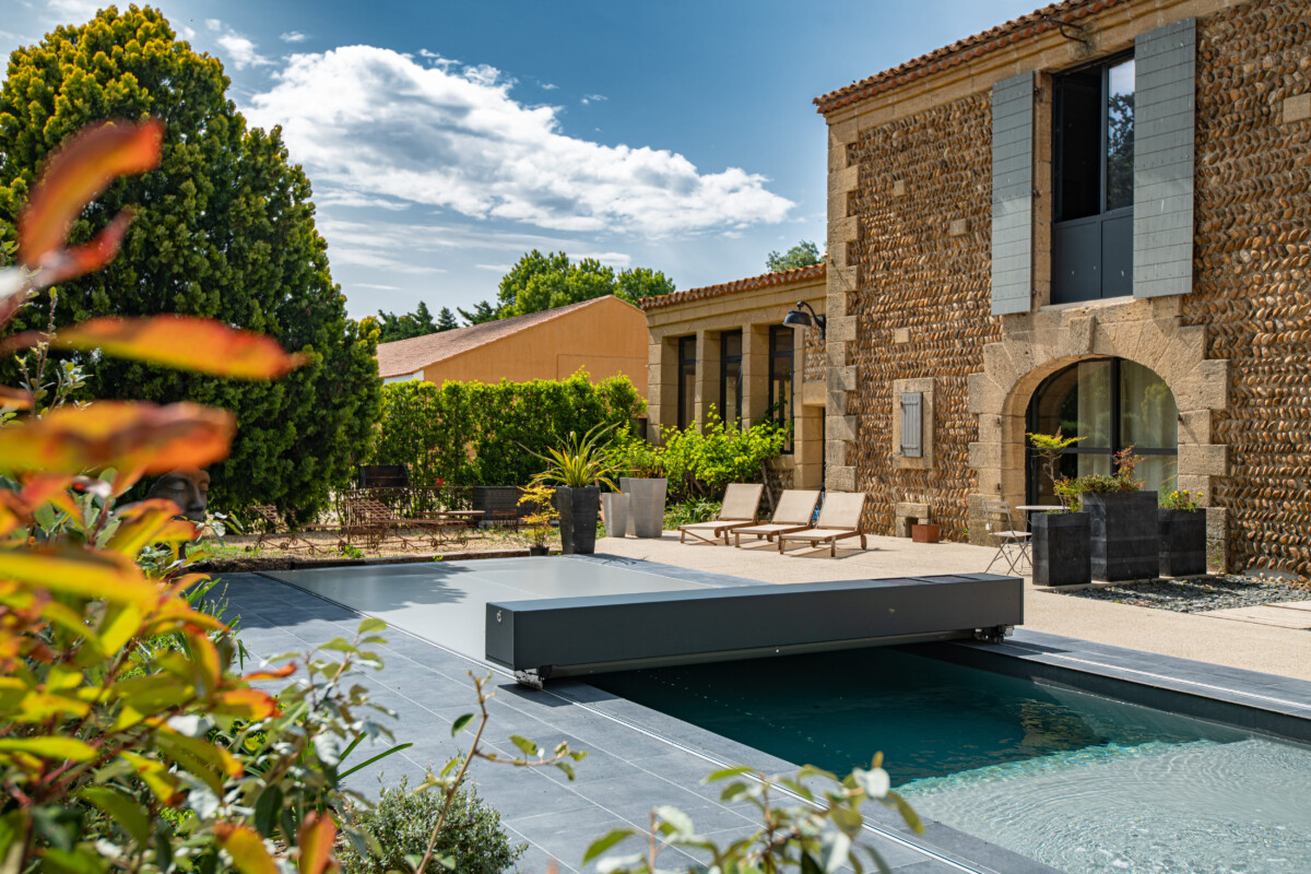 Maison en pierre avec volets gris, piscine couverte, transats et végétation environnante sous un ciel partiellement nuageux.