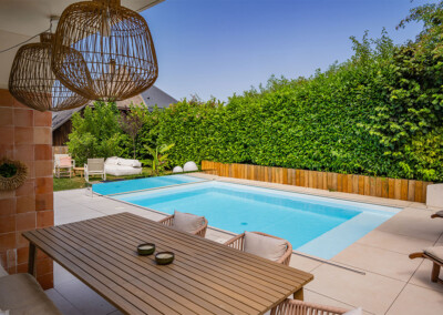 Coverseal-landscape7 Vue depuis une terrasse couverte donnant sur une piscine équipée d’une Coverseal, entourée de haies végétales et d’un mobilier de jardin naturel.